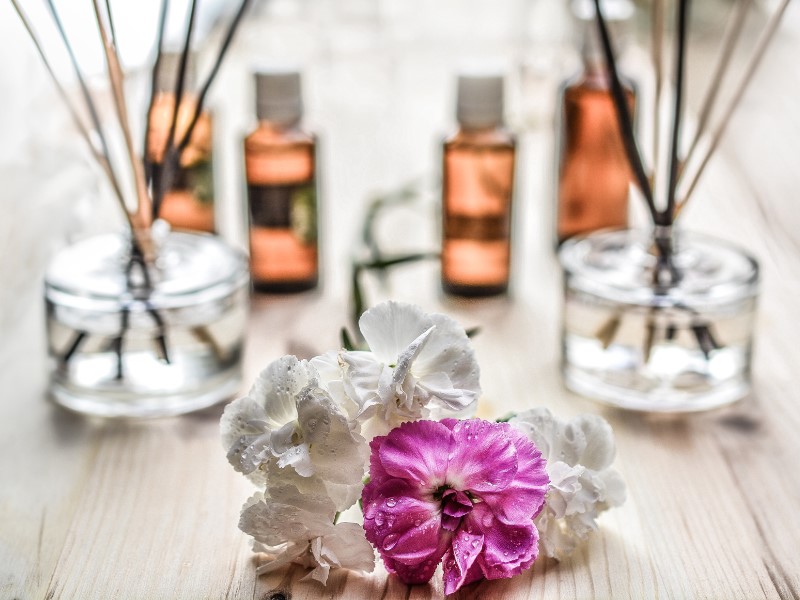 Image shows pretty flower in shades of purple surrounded by white flowers all with water drops placed on a wooden surface with four bottle scents flanked by two glass jars filled with scent sticks blurred in the background.