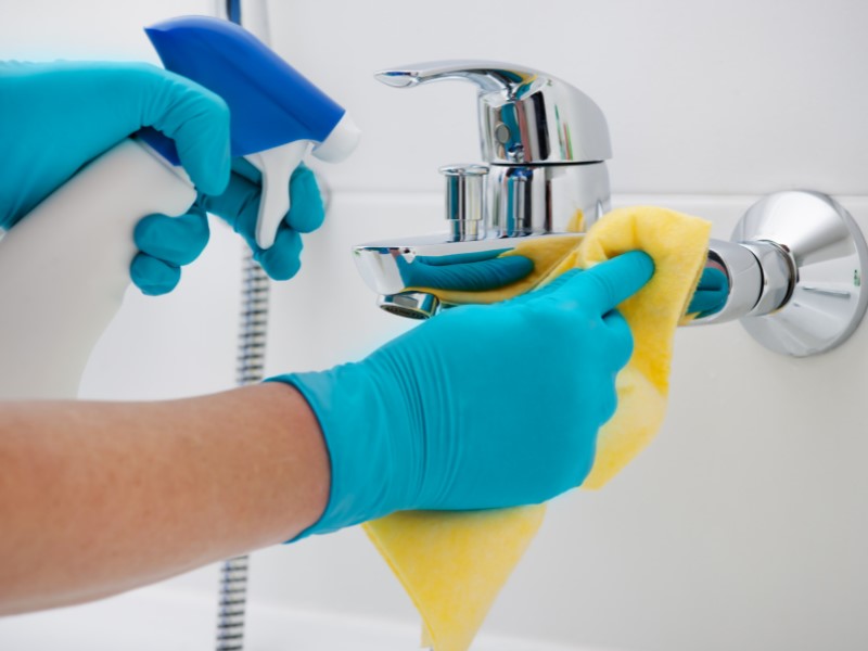 Image shows blue gloved hands cleaning a silver tap in what looks like a white tiled bathroom with a spray bottle and a yellow wash rag.