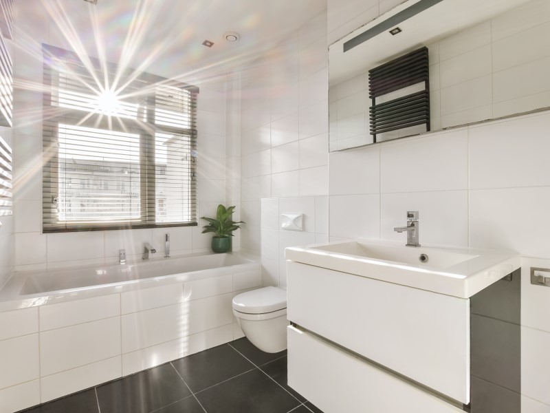 Image shows a small aesthetically pleasant white tile bathroom with a white bathtub, sink and toilet, silver accented with the taps as well as a well placed white ventilation system and a green potted house plant strategically placed beneath a big wooden paned window with blinds letting in some gorgeous sunshine.